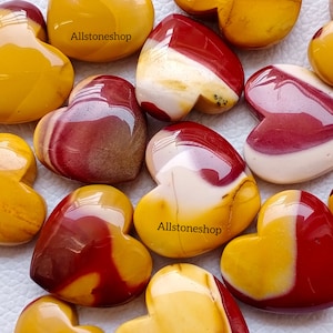 May include: A collection of heart-shaped polished stones in shades of red, yellow, and white. The stones are arranged in a random pattern. The text "Allstoneshop" appears twice in the image.