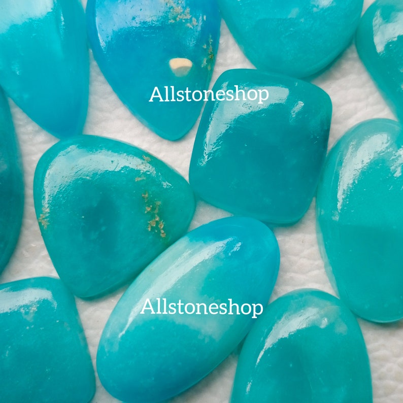 Puede incluir: Una colecci&oacute;n de ocho cabujones turquesa de diferentes formas y tama&ntilde;os. Los cabujones son lisos y pulidos, de color azul claro y con un ligero brillo. Los cabujones est&aacute;n dispuestos sobre una superficie blanca.