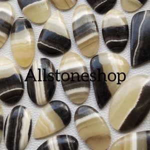 May include: A collection of 20+ black and white banded agate cabochons. The cabochons are oval and teardrop shaped. The banding is natural and varies in width and color. Allstoneshop is printed on the white background.