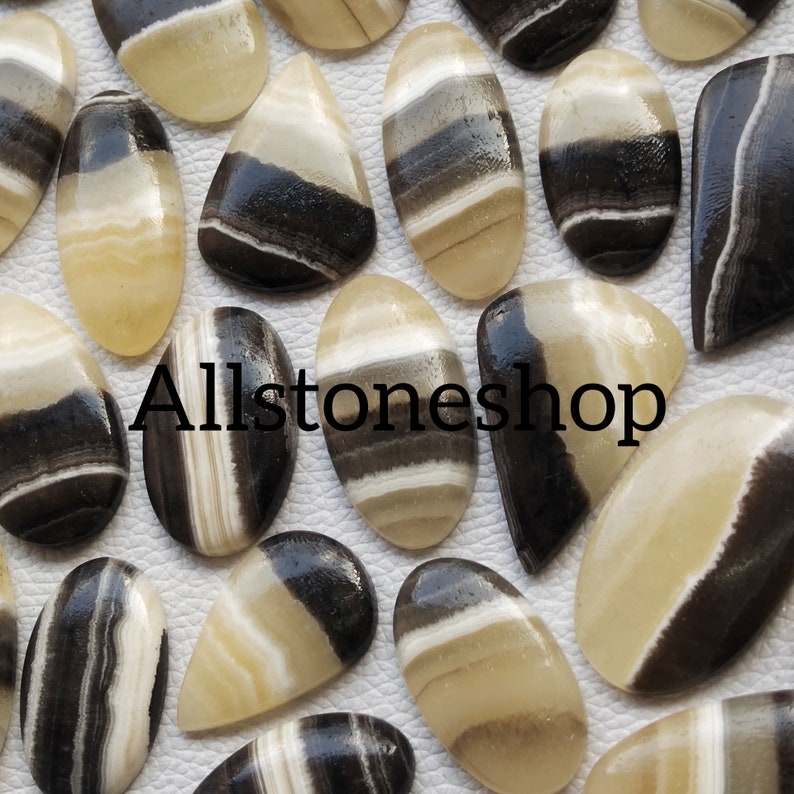 May include: A collection of 20+ black and white banded agate cabochons. The cabochons are oval and teardrop shaped. The banding is natural and varies in width and color. Allstoneshop is printed on the white background.