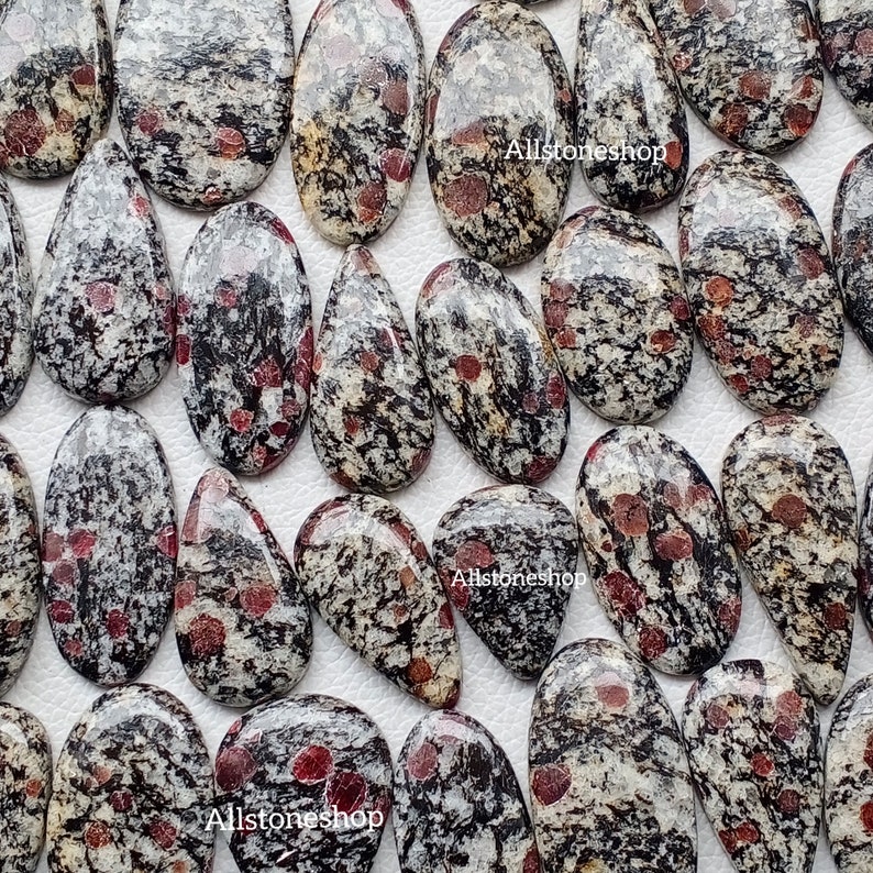May include: A selection of polished gemstones with a speckled pattern of black, white, and red. The stones are oval and teardrop-shaped, with a smooth, glossy finish. The image includes the text "Allstoneshop".