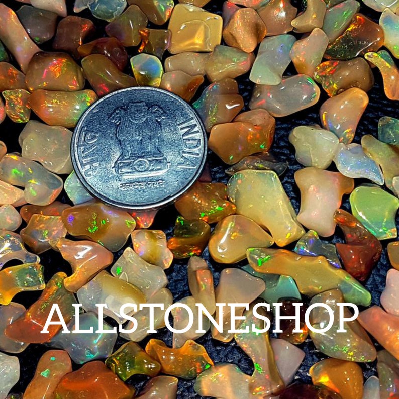 May include: A close-up of a pile of small, iridescent opal chips in various shades of orange, yellow, and white. The chips are scattered on a dark surface, and a silver Indian coin is visible in the center of the image. The text "ALLSTONESHOP" is visible at the bottom of the image.
