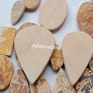 May include: A collection of natural stone cabochons in various shapes and sizes. The stones are a light beige color with brown and yellow markings. The stones are arranged on a white surface. Allstoneshop is printed on the image.