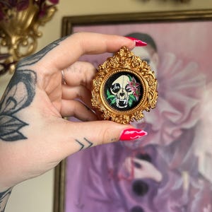 May include: A small, ornate gold-colored frame with a black background, featuring a skull with pink flowers and green leaves. The frame is held in a hand with a tattoo. The background includes a framed painting and decorative elements.