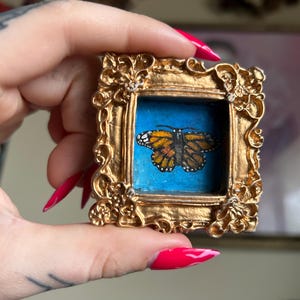 May include: A miniature painting of a monarch butterfly in a gold ornate frame. The butterfly has orange, black, and white markings against a blue background. The frame is highly detailed with floral and scroll designs.
