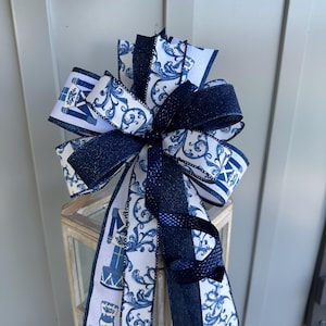 May include: A decorative bow featuring navy blue and white ribbons. The bow has a layered design with ribbons displaying a swirl pattern and a nutcracker soldier motif. The bow is attached to a small, rustic, wooden lantern.