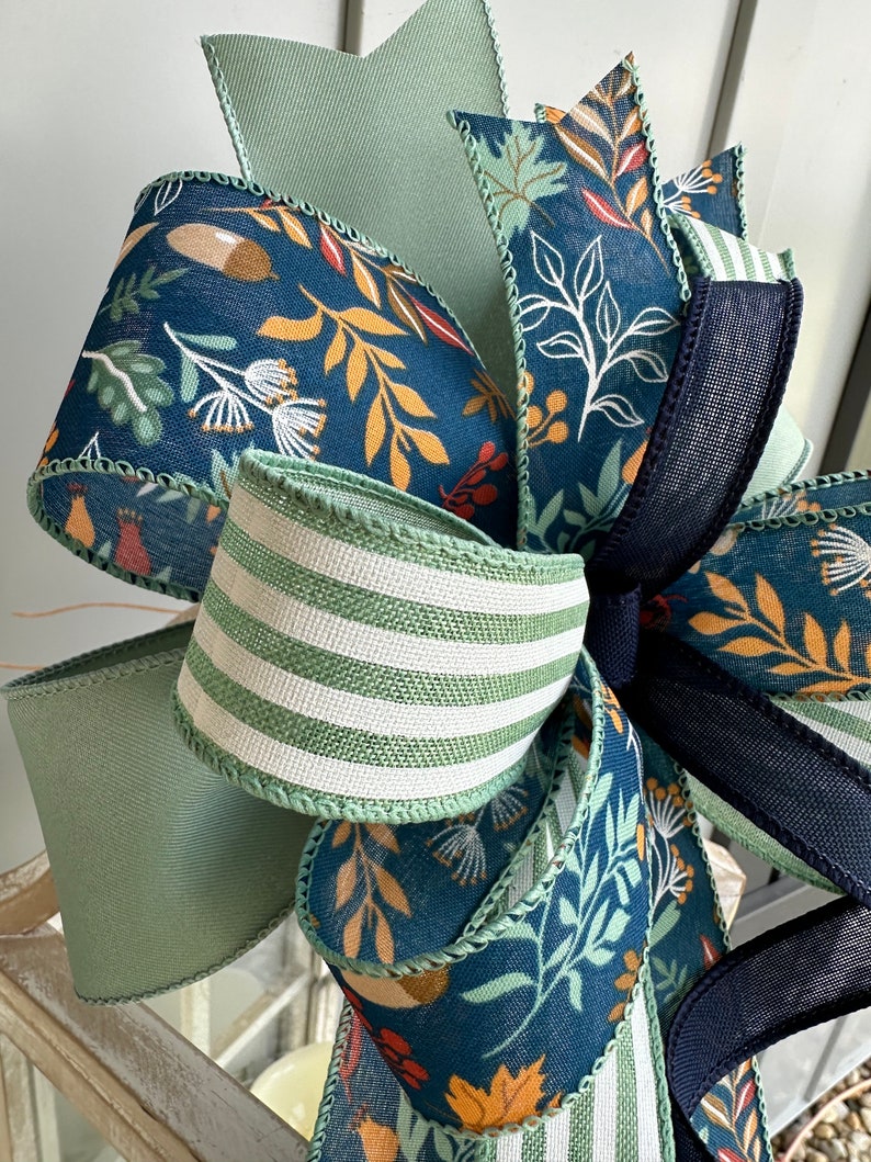 May include: A decorative bow made with three different ribbons. The ribbons are a blue and white floral print, a green and white striped ribbon, and a solid green ribbon. The bow is tied with a black ribbon.