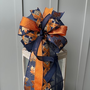 May include: A navy blue and orange plaid ribbon bow with gold leaf accents. The bow is tied around a lantern.