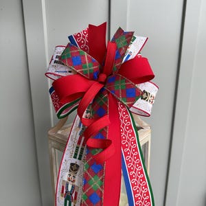 May include: A festive Christmas bow featuring a large red bow with long, flowing ribbons. The bow incorporates patterned ribbons with nutcracker soldiers, plaid designs, and white scrollwork. Accents include green, blue, and gold.
