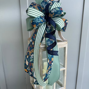 May include: A decorative bow made with three different ribbons. The bow is tied around a white lantern. The ribbons are green with white stripes, green with a floral pattern, and navy blue with a floral pattern.