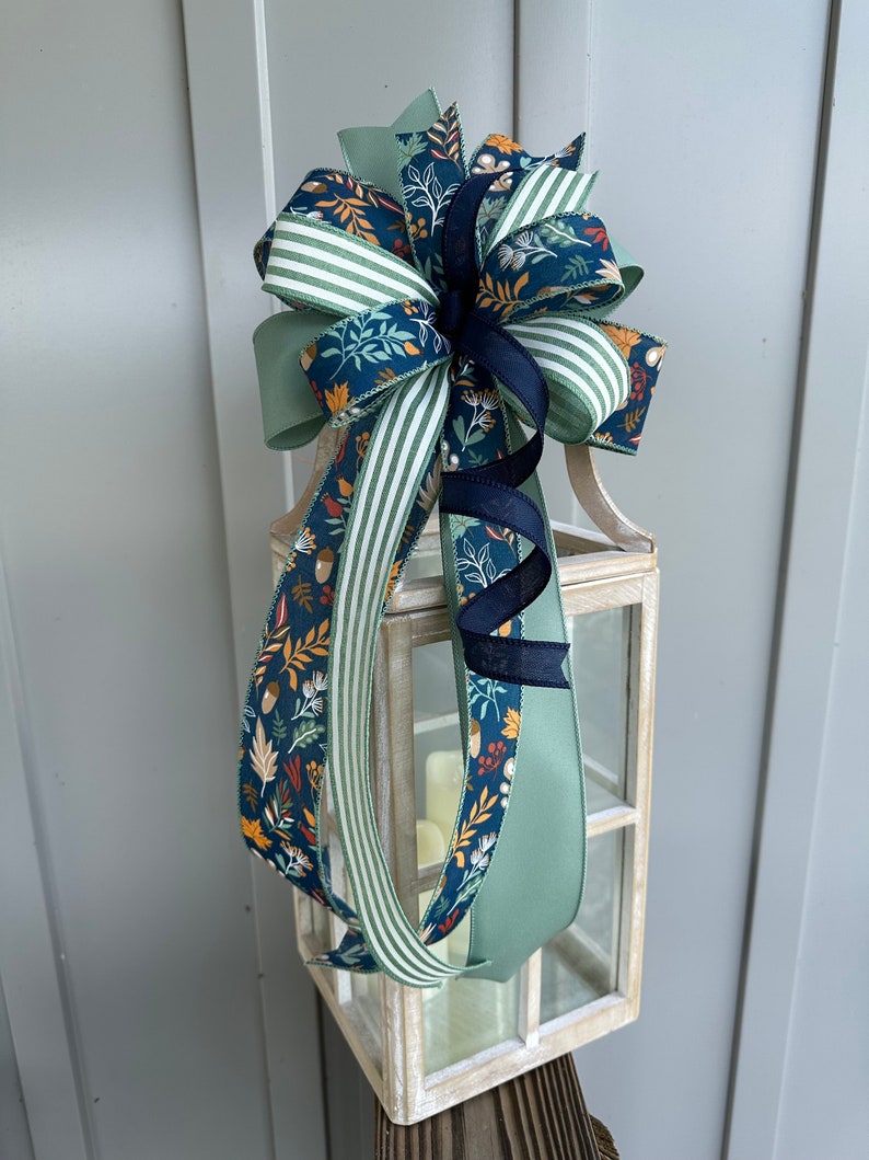 May include: A decorative bow made with three different ribbons. The bow is tied around a white lantern. The ribbons are green with white stripes, green with a floral pattern, and navy blue with a floral pattern.