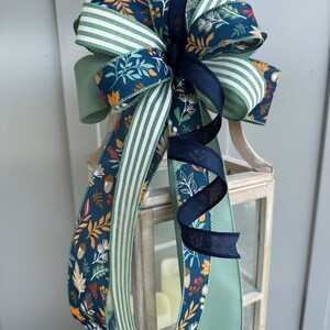 May include: A decorative bow made with three different ribbons. The ribbons are green with white stripes, green with a floral pattern, and navy blue with a floral pattern. The bow is tied with a black ribbon.