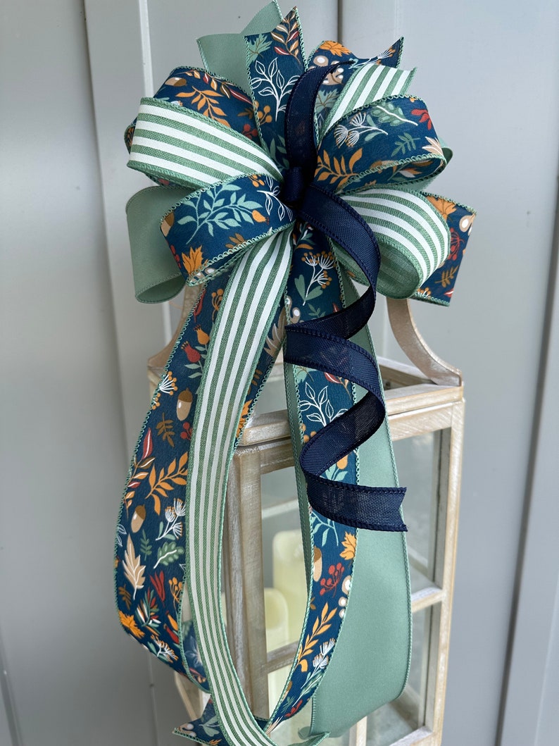 May include: A decorative bow made with three different ribbons. The ribbons are green with white stripes, green with a floral pattern, and navy blue with a floral pattern. The bow is tied with a black ribbon.