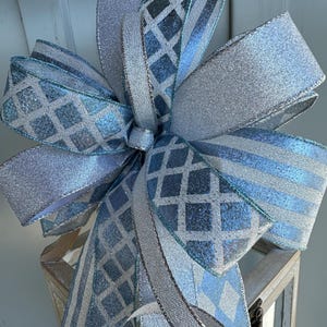 May include: A decorative bow crafted from multiple ribbons featuring a shimmering silver glitter finish, a light blue and silver diamond lattice pattern, and light blue and silver stripes with wired edges for structure and volume.