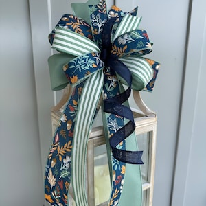 May include: A decorative bow made with three different ribbons. The ribbons are a green and white striped ribbon, a solid green ribbon, and a blue ribbon with a floral pattern in shades of orange, red, and yellow. The bow is tied around a lantern.