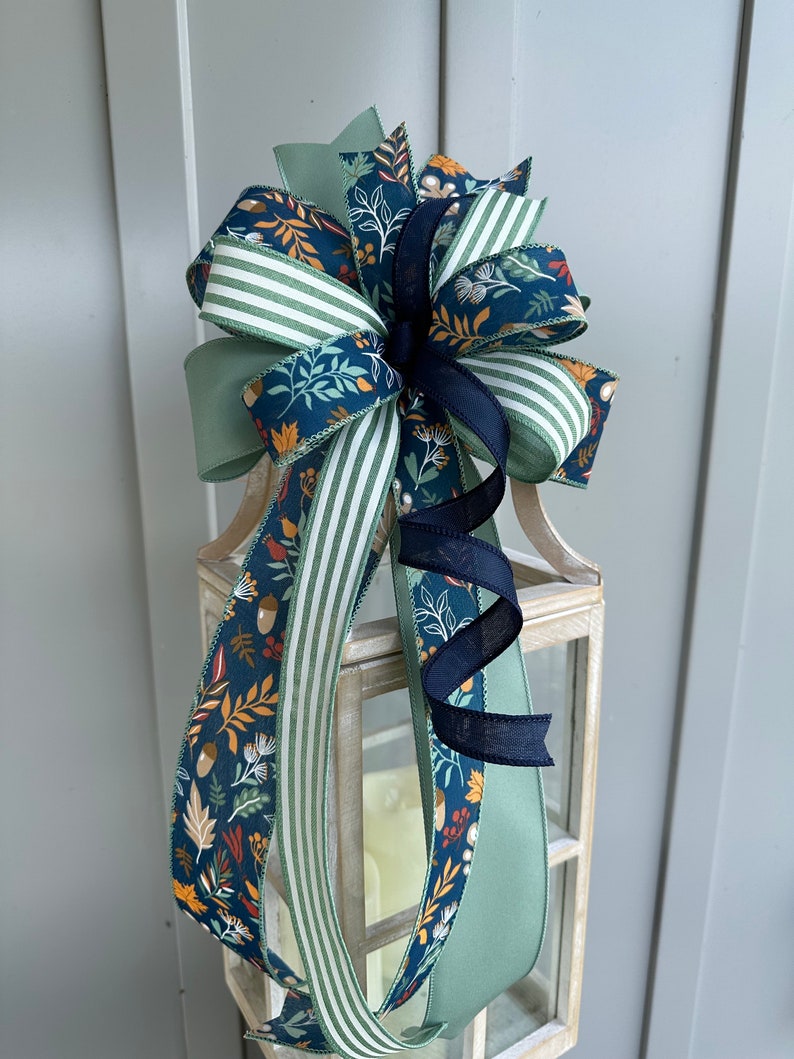 May include: A decorative bow made with three different ribbons. The ribbons are a green and white striped ribbon, a solid green ribbon, and a blue ribbon with a floral pattern in shades of orange, red, and yellow. The bow is tied around a lantern.