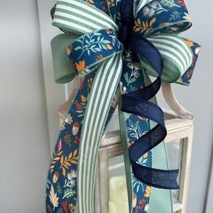 May include: A festive bow made with three different ribbons. The ribbons are a green and white striped ribbon, a solid sage green ribbon, and a blue ribbon with a fall-themed print of leaves, acorns, and berries.