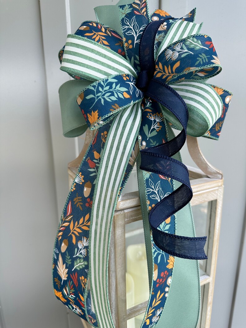 May include: A festive bow made with three different ribbons. The ribbons are a green and white striped ribbon, a solid sage green ribbon, and a blue ribbon with a fall-themed print of leaves, acorns, and berries.