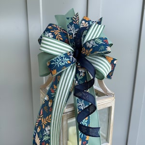 May include: A decorative bow made with three different ribbons. The ribbons are green, white, and blue with a pattern of leaves, acorns, and stripes. The bow is tied around a lantern.