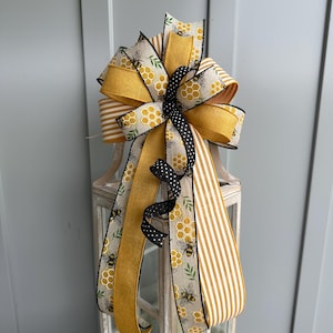May include: A decorative bow featuring a bee theme. The bow has multiple layers of ribbon in yellow, black, and white. Some ribbons have a honeycomb and bee pattern, while others are striped or solid. The bow is attached to a white lantern.