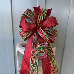 May include: A large Christmas bow with a red glitter finish and striped green and red ribbon accents. The bow is tied and has long, flowing tails, ideal for festive decorations.