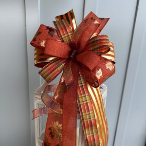 May include: A large, decorative bow made of three different ribbons. The ribbons are a rust color with gold stripes, a plaid pattern with rust, gold, and green, and a rust color with gold leaf accents. The bow is tied around a lantern.