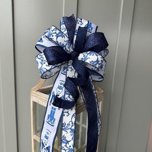 May include: A decorative bow crafted from multiple ribbons, featuring a white background with blue nutcracker figures and intricate blue scrollwork patterns. The design includes solid dark navy blue glittery ribbon accents.