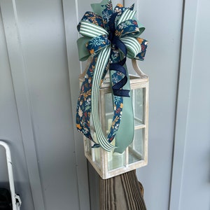 May include: A white lantern with a green, white and blue ribbon bow on top. The lantern is sitting on a wooden post.