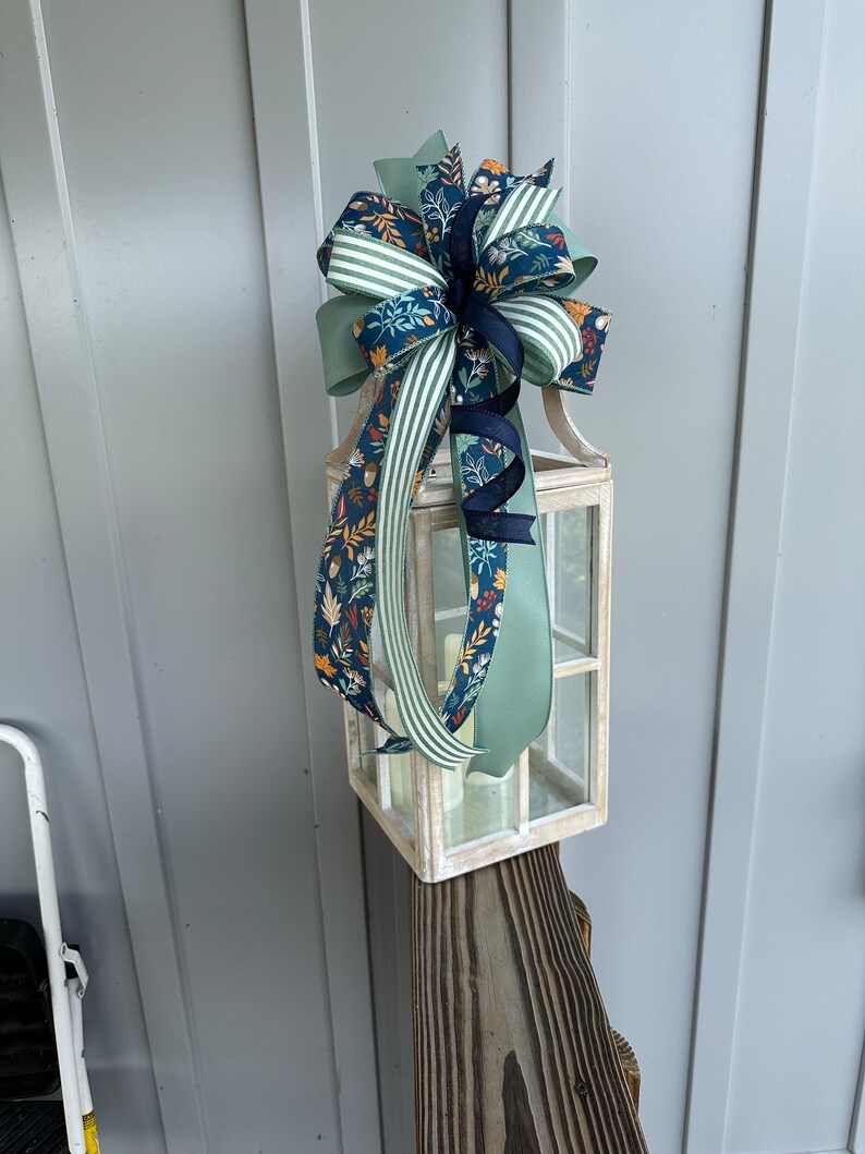 May include: A white lantern with a green, white and blue ribbon bow on top. The lantern is sitting on a wooden post.