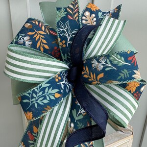 May include: A large, decorative bow made with multiple ribbons in shades of green, blue, and brown. The ribbons feature a variety of autumn-themed patterns, including leaves, acorns, and berries. The bow is tied with a black ribbon.