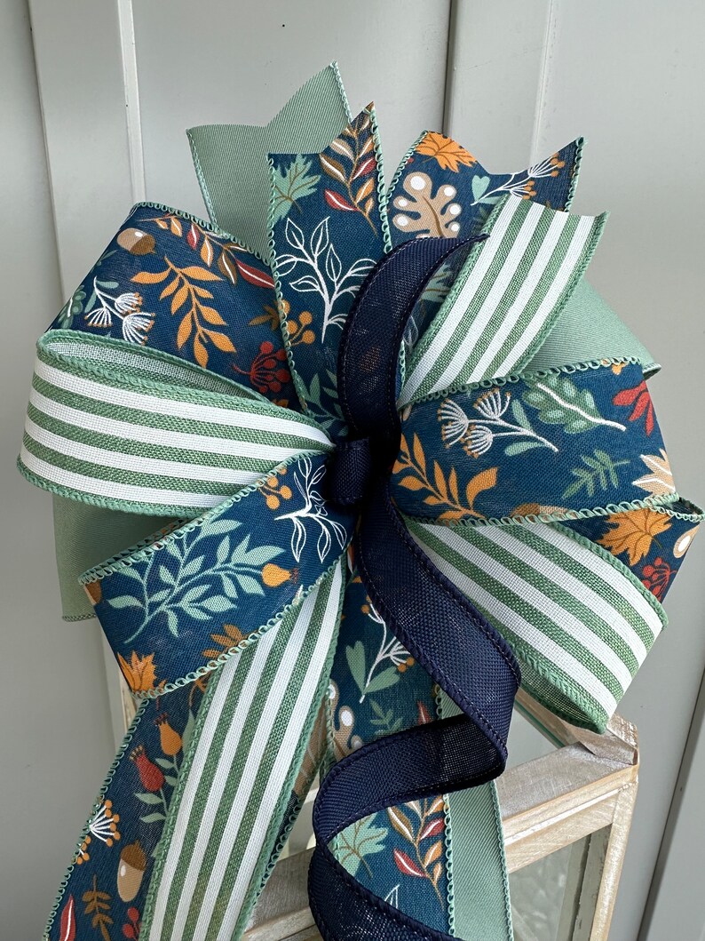 May include: A large, decorative bow made with multiple ribbons in shades of green, blue, and brown. The ribbons feature a variety of autumn-themed patterns, including leaves, acorns, and berries. The bow is tied with a black ribbon.