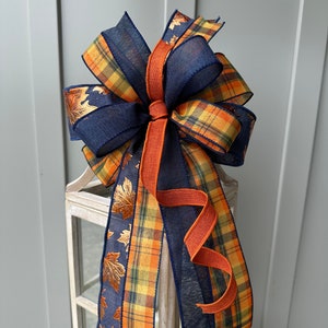 May include: A fall-themed bow made with navy blue, orange, and yellow plaid ribbon, and a gold leaf ribbon. The bow is tied with a rust-colored ribbon.