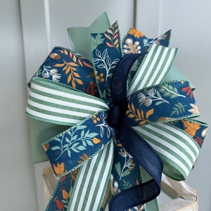 May include: A green, white and navy blue bow with a floral print. The bow is made of ribbon and has a wired edge.