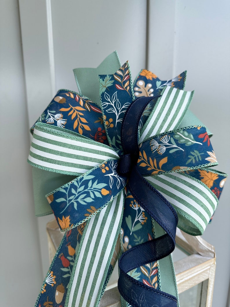 May include: A green, white and navy blue bow with a floral print. The bow is made of ribbon and has a wired edge.