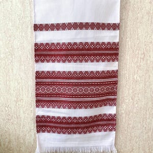 Könnte beinhalten: Weißes rechteckiges Textil mit aufwendigen roten Stickmustern. Das Design zeigt horizontale Bänder mit geometrischen und floralen Motiven. Der untere Rand hat Fransen. Dieses dekorative Stück könnte ein Handtuch oder ein Tischläufer sein.