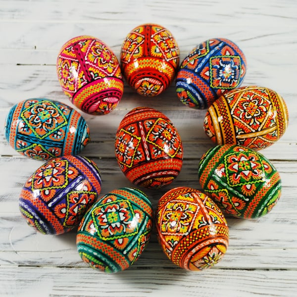 May include: A collection of vibrantly decorated Easter eggs. Each egg features intricate geometric and floral patterns in a variety of colors, including red, orange, blue, green, and yellow. The eggs are arranged on a white wooden surface.