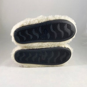 Puede incluir: La parte inferior de un par de zapatillas blancas y peludas con suelas de goma negra. Las suelas tienen un patr&oacute;n ondulado.