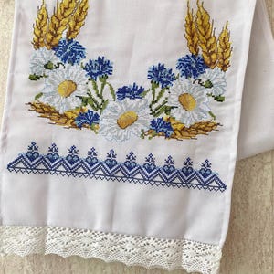 Könnte beinhalten: Weißes besticktes Handtuch mit einem floralen Design aus blauen Kornblumen, weißen Gänseblümchen und goldenem Weizen. Der untere Rand hat ein blaues geometrisches Muster und eine weiße Spitzenborte. Ein dekoratives Küchen- oder Badtextil.