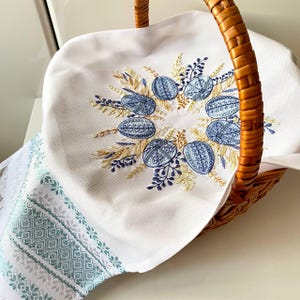 May include: A woven basket with a white cloth featuring embroidered blue Easter eggs and floral accents. The cloth has a decorative border with a geometric pattern in shades of blue and green. The basket handle is made of woven brown material.