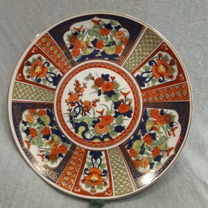 Puede incluir: Un plato de cerámica blanca con un borde dorado y un diseño floral colorido. El diseño presenta flores y hojas rojas, naranjas, azules y verdes. El plato está decorado con un patrón geométrico en el fondo.