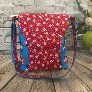 Puede incluir: Un bolso bandolera de tela rojo y azul con cierre de solapa. El bolso presenta un estampado floral y una imagen de dibujos animados de Mickey Mouse y Minnie Mouse.