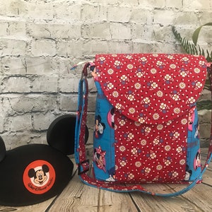 Puede incluir: Un bolso bandolera de tela azul y rojo con estampado floral y cierre de solapa. El bolso presenta un dise&ntilde;o de Mickey Mouse y Minnie Mouse. El bolso est&aacute; sobre una superficie de madera con un gorro de orejas de Mickey Mouse negro.