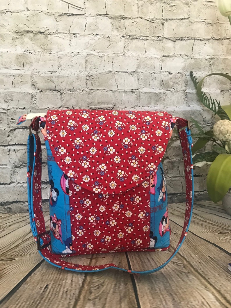 Puede incluir: Un bolso bandolera de tela rojo y azul con cierre de solapa. El bolso presenta un estampado floral y una imagen de dibujos animados de Mickey Mouse y Minnie Mouse.