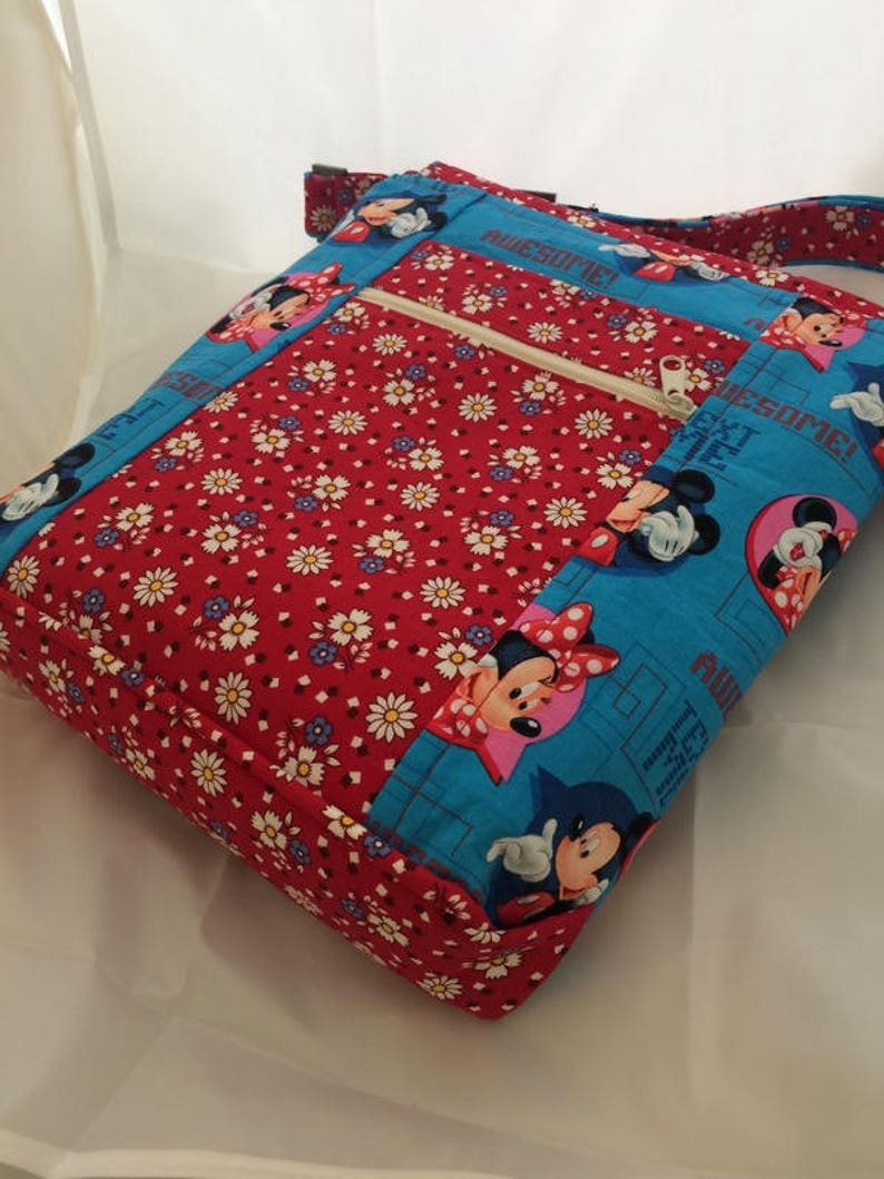 Puede incluir: Un bolso bandolera de tela rojo y azul con un bolsillo con cremallera y un estampado de Mickey Mouse y Minnie Mouse. El bolso tiene un estampado de margaritas blancas en la solapa delantera y las palabras "Awesome!" impresas en el lateral.
