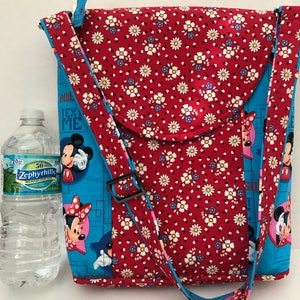 Puede incluir: Bolso de tela rojo y azul con estampado floral y correa negra. El bolso tiene una solapa con borde redondeado. El bolso presenta un dise&ntilde;o de Mickey Mouse y Minnie Mouse con el texto "Awe, Test Me".