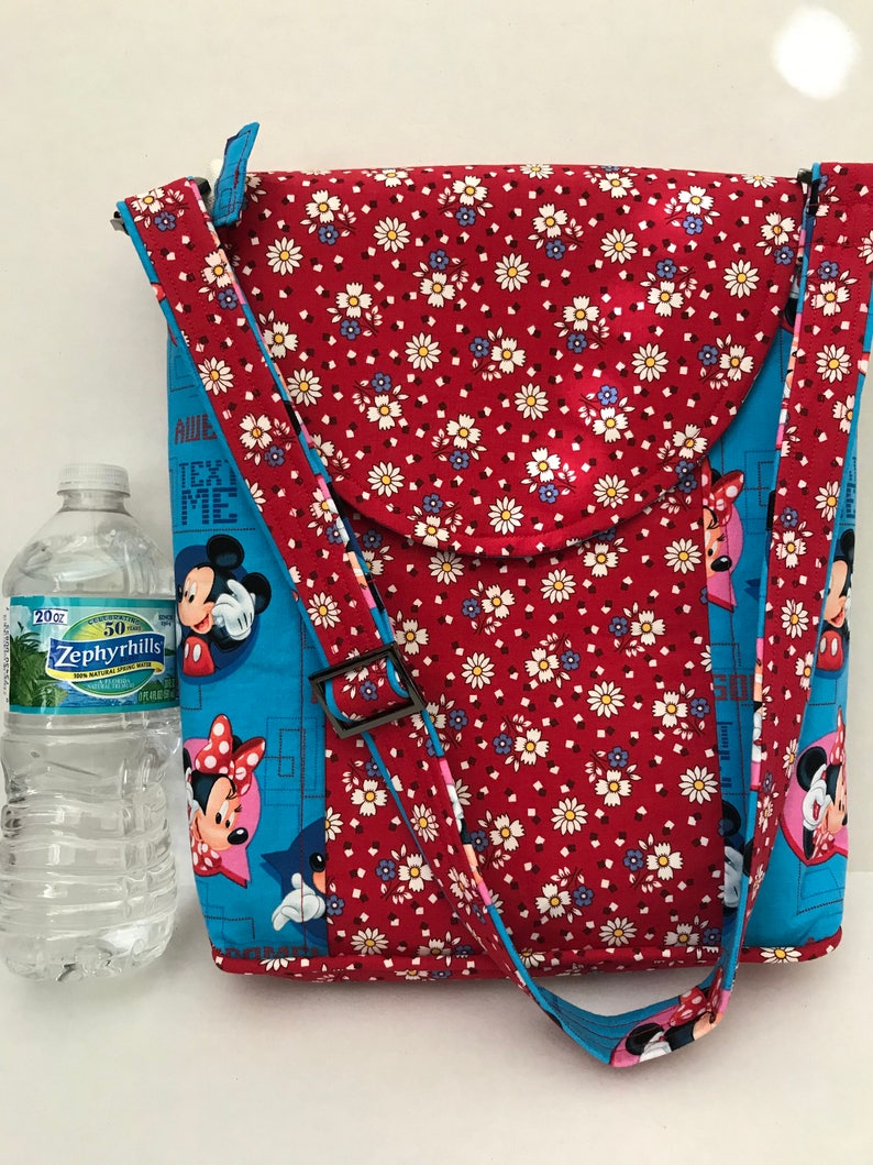 Puede incluir: Bolso de tela rojo y azul con estampado floral y correa negra. El bolso tiene una solapa con borde redondeado. El bolso presenta un dise&ntilde;o de Mickey Mouse y Minnie Mouse con el texto "Awe, Test Me".