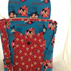 Puede incluir: Un bolso bandolera de tela azul y roja con solapa superior. El bolso presenta un estampado floral y un estampado de Mickey Mouse y Minnie Mouse. La solapa tiene el texto "Awesome! Text Me".