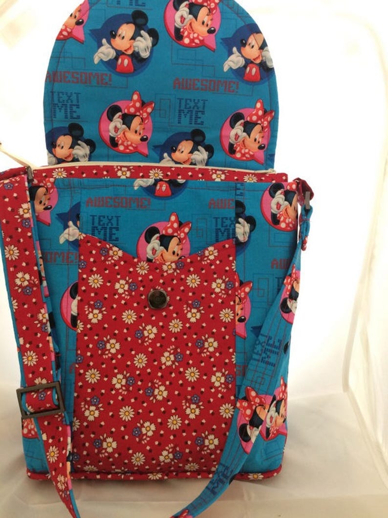 Puede incluir: Un bolso bandolera de tela azul y roja con solapa superior. El bolso presenta un estampado floral y un estampado de Mickey Mouse y Minnie Mouse. La solapa tiene el texto "Awesome! Text Me".