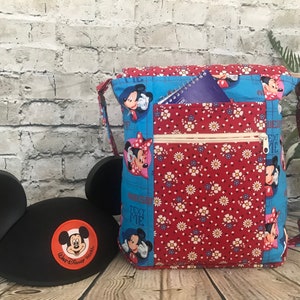 Puede incluir: Una bolsa de tela azul y roja con un bolsillo con cremallera y un estampado de Mickey Mouse y Minnie Mouse. La bolsa tiene una correa larga y un peque&ntilde;o bolsillo en la parte delantera. El texto "Text Me" est&aacute; impreso en la bolsa.