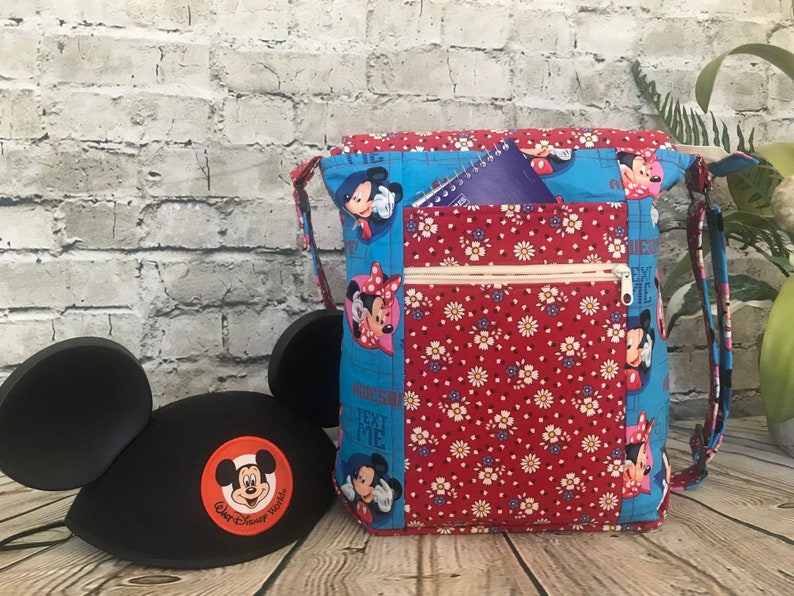 Puede incluir: Una bolsa de tela azul y roja con un bolsillo con cremallera y un estampado de Mickey Mouse y Minnie Mouse. La bolsa tiene una correa larga y un peque&ntilde;o bolsillo en la parte delantera. El texto "Text Me" est&aacute; impreso en la bolsa.